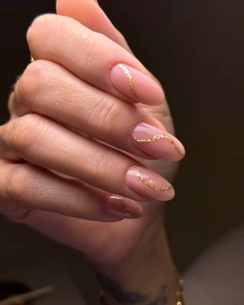 Close-up of a hand with glossy pink acrylic nails in an almond shape, decorated with curved gold glitter accents against a softly blurred background