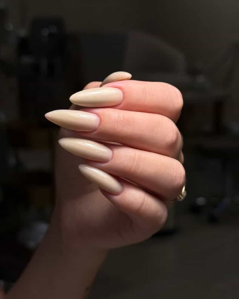 A hand with long almond-shaped acrylic nails painted in a glossy nude beige shade, held under soft salon lighting