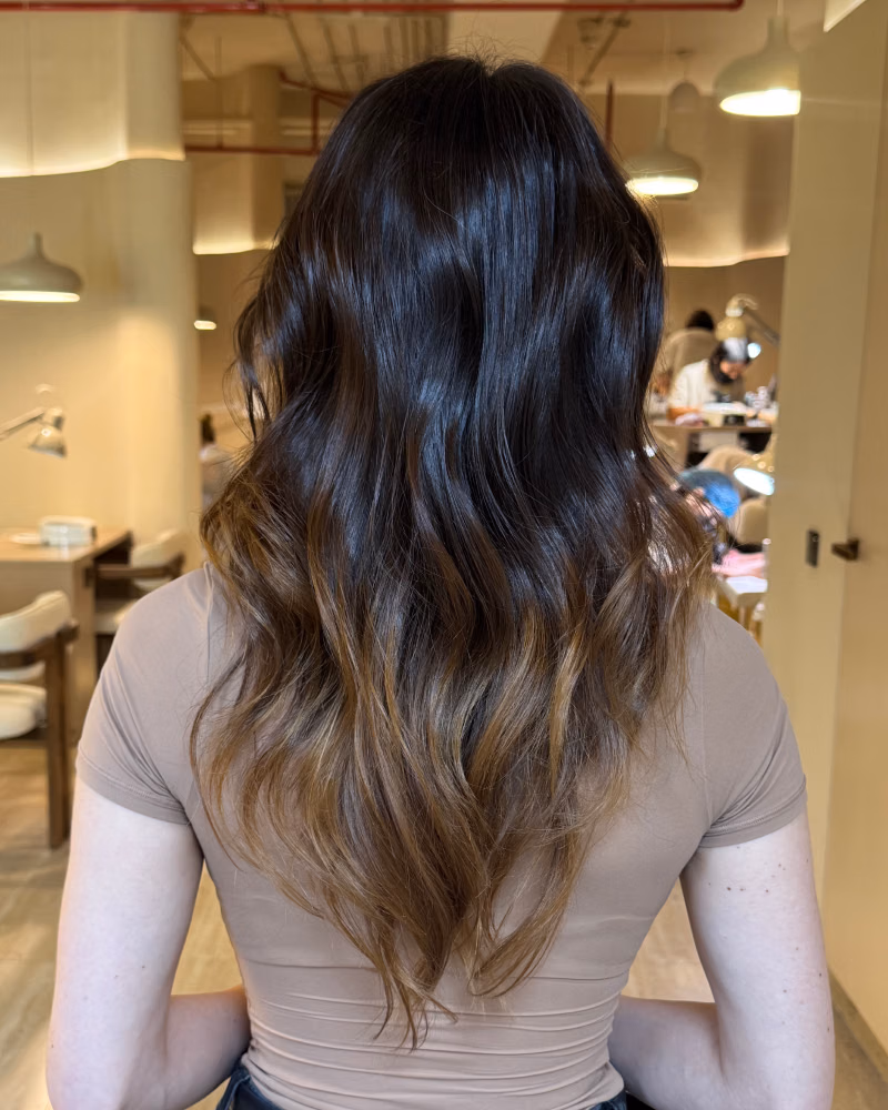Back view of a woman with long wavy brunette hair featuring warm caramel balayage ends, sitting in a softly lit salon setting