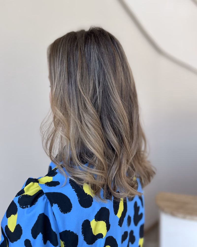 Back view of a woman with shoulder-length wavy balayage hair, featuring dark roots and soft blonde highlights, wearing a bright blue patterned top