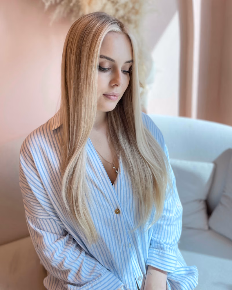 A blonde woman with soft balayage hair sits on a pale sofa, looking down, wearing a light blue striped shirt against a soft pink interior backdrop