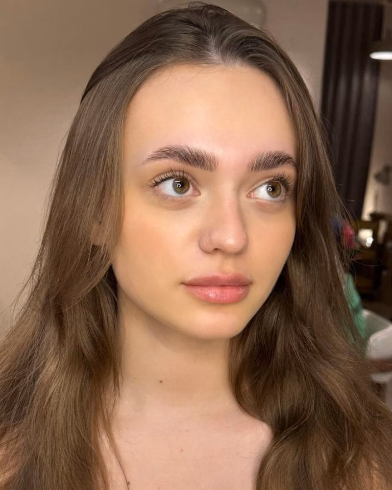 Woman with long brown hair and softly glowing makeup showcasing freshly shaped brows after brow threading and lamination