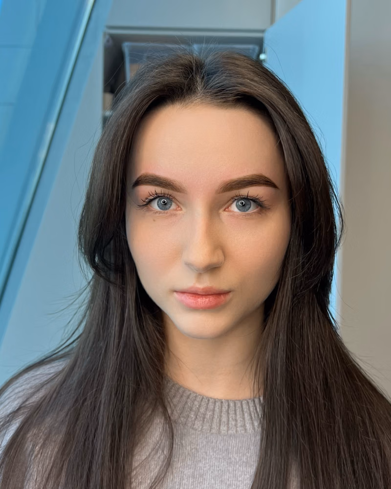 a woman with long dark hair, blue eyes, shaped brows, and subtle eyelash extensions, wearing a light grey sweater indoors near a window