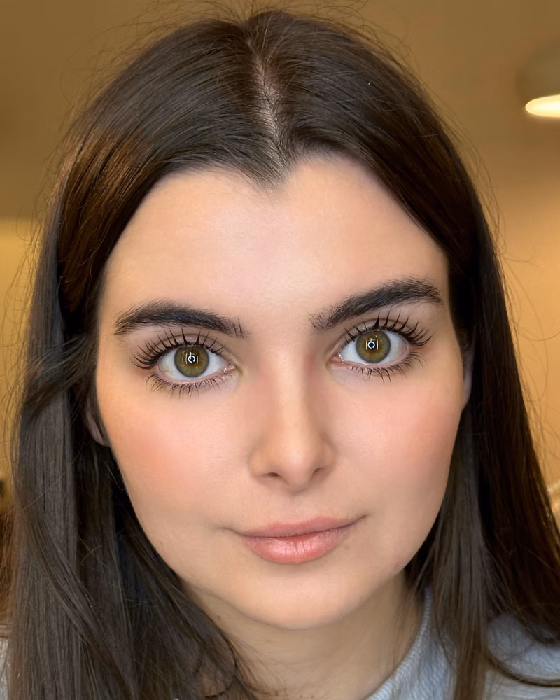 a woman with long dark brown hair, green eyes, soft makeup, and visible eyelash extensions, wearing a grey sweatshirt