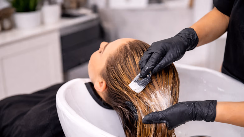 Hairdresser applying hair color treatment to a client’s hair at a salon wash basin using protective gloves