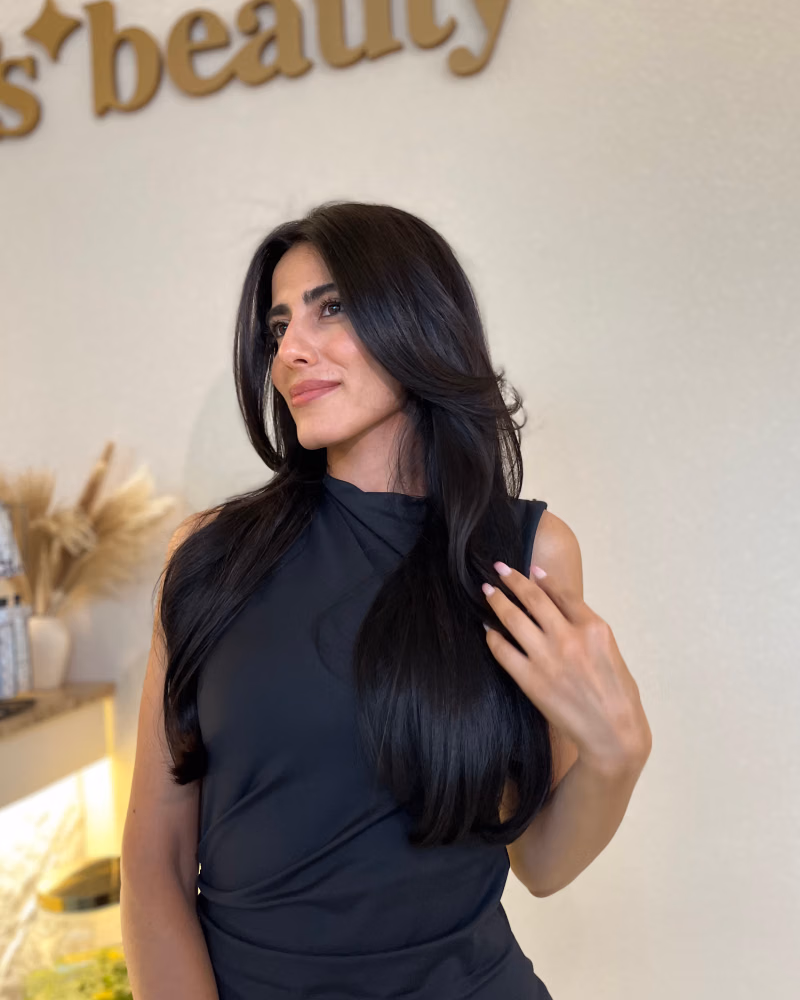 Woman with long, glossy dark brunette hair styled in soft face-framing layers, posing in a black sleeveless top inside a beauty salon
