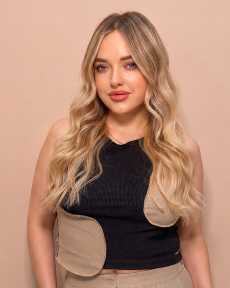 Woman with long wavy blonde hair and soft special colouring poses against a pale pink background, wearing a black and beige top