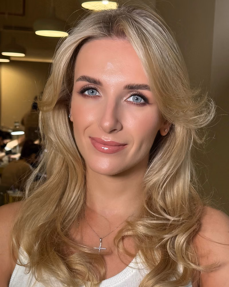 Blonde woman with soft curled hair posing indoors, wearing glowing glam makeup with defined brows, fluttery lashes, rosy cheeks, and glossy pink lips