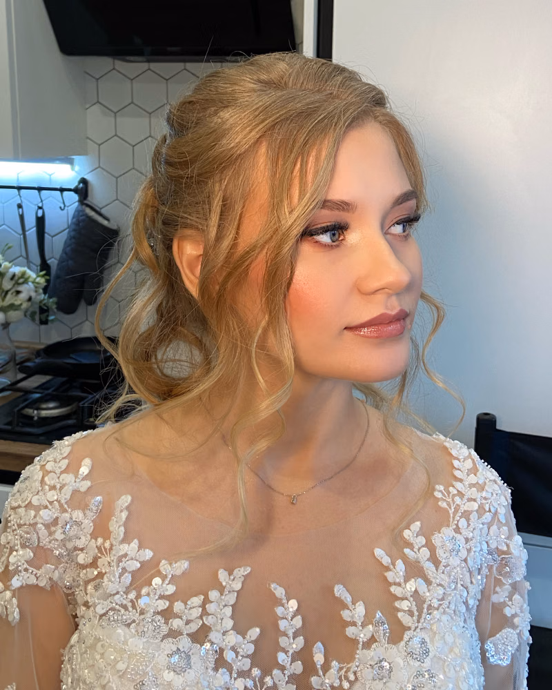 Blonde woman in an embellished white bridal-style dress, wearing soft romantic makeup with defined lashes, warm eyeshadow, rosy blush, glowing skin, and glossy nude-pink lips