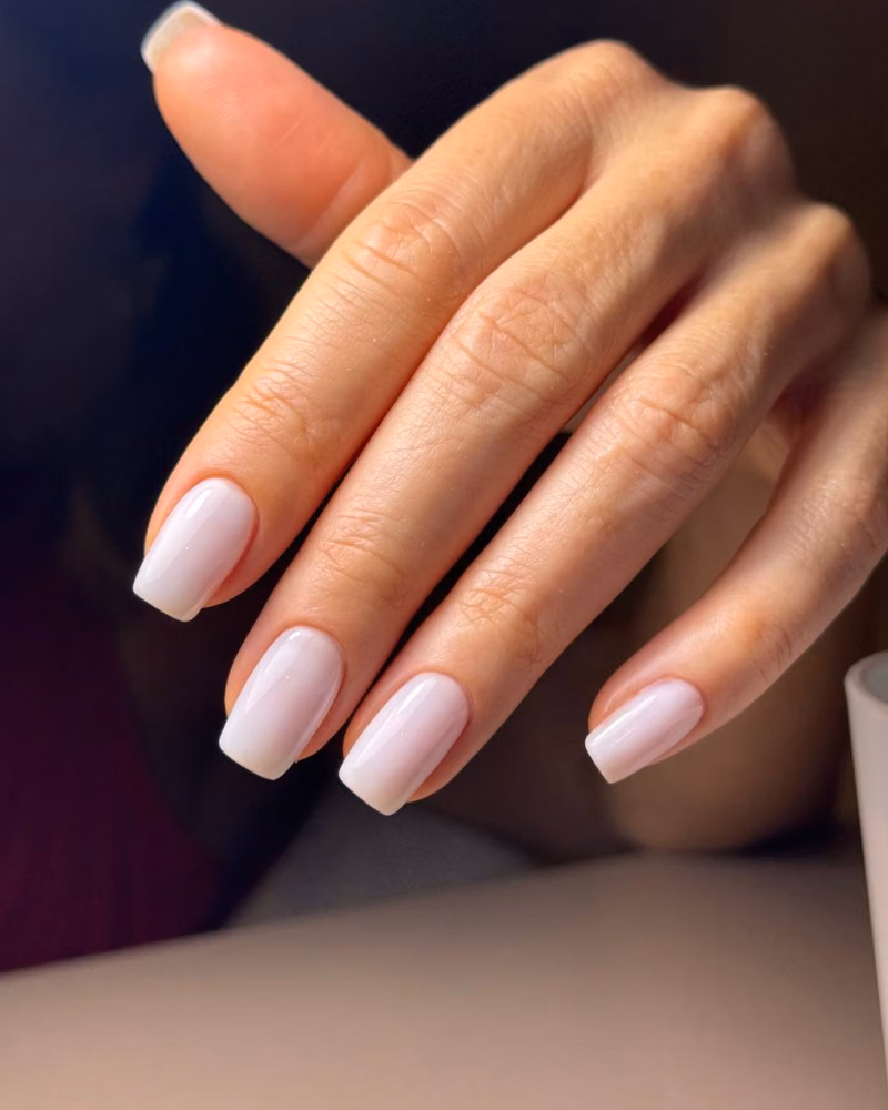 a hand with glossy pale pink gel nails in a soft square shape, shown in warm natural light