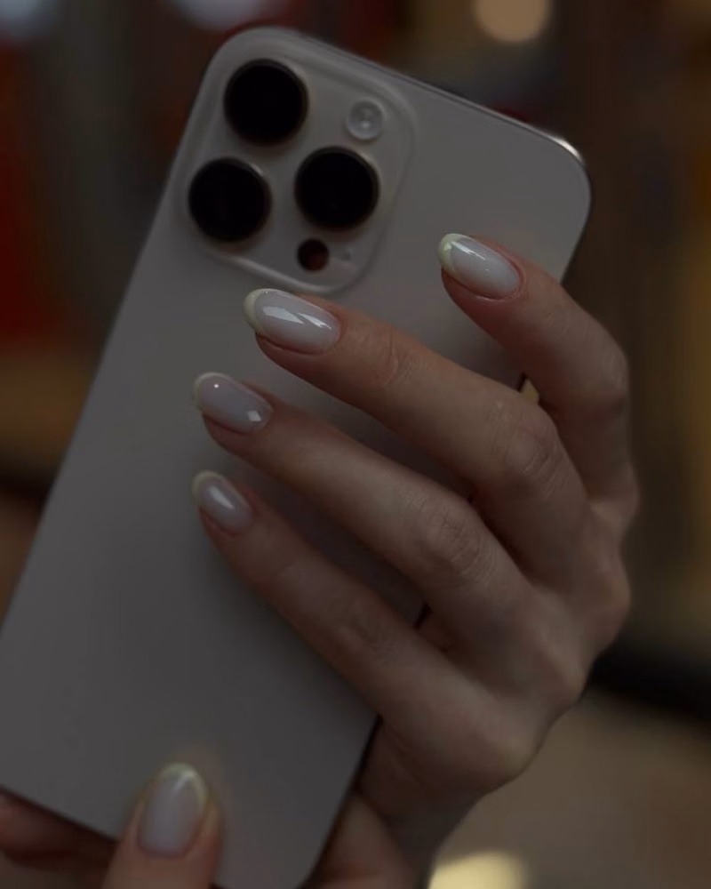 a hand holding a silver iPhone, showing glossy nude gel nails with soft white tips