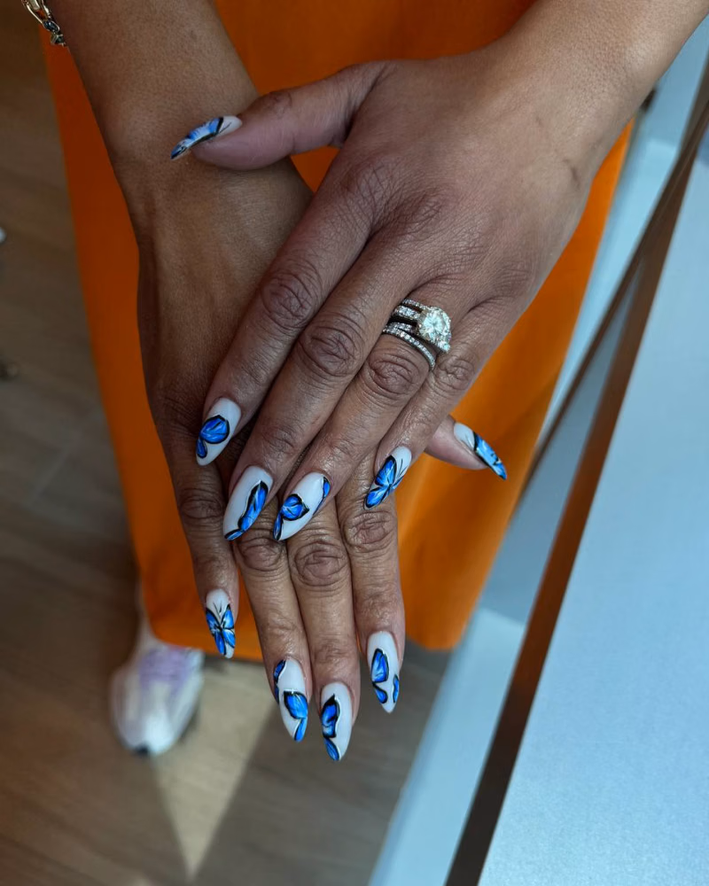 Hands showing long almond-shaped nails with a white base and detailed blue butterfly nail art design, accented with black outlines and glossy finish