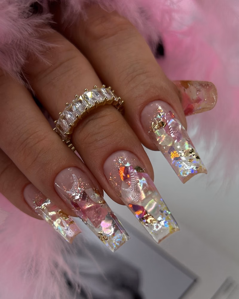 Long square acrylic nails with a nude pink base and glittery encapsulated nail art design featuring iridescent flakes, gold foil, and reflective accents