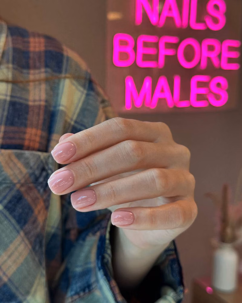 A hand showing glossy pale pink manicured nails in front of a neon “Nails Before Males” sign
