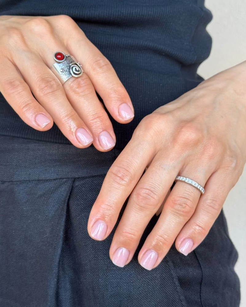 A hand showing glossy pale pink manicured nails in front of a neon “Nails Before Males” sign