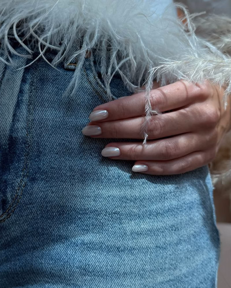 a hand with a pearly Russian manicure resting on light blue denim, framed by soft white feather details