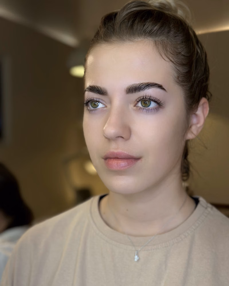 woman with neatly styled brows and Russian volume lashes, wearing a beige top and delicate necklace in soft indoor lighting