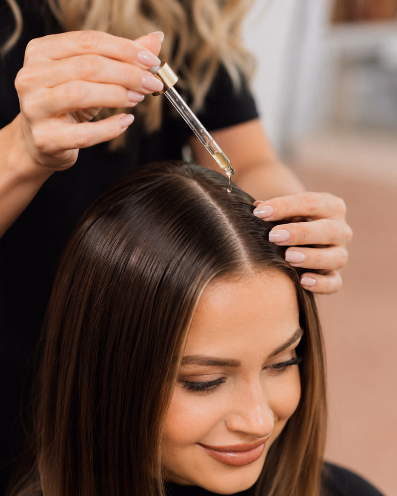Hairstylist applying hair serum with a glass dropper to a woman’s center part during a salon scalp treatment